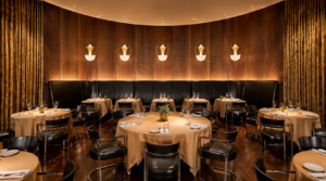 Elegant dining room with round beige-tablecloth tables, black chairs, and warm amber lighting along wooden-paneled walls