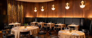Elegant restaurant dining room with beige-tablecloth tables, black chairs, and warm pendant lights along a wood-paneled wall.