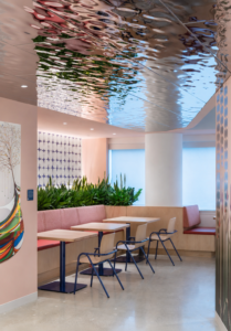 Modern cafe seating: pink banquette, light wood tables, and blue chairs with green plants as a divider.