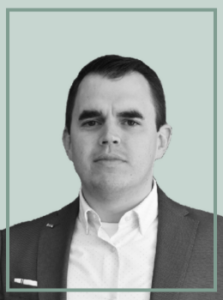 Professional headshot of a man in a dark suit and white shirt, looking at the camera against a pale teal background.