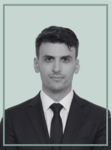 Professional headshot of a man in a dark suit and tie, neutral expression, light teal background.