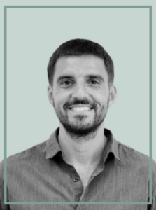 Portrait of a smiling man with short dark hair and a beard, wearing a gray button-down shirt against a light teal background, head and shoulders.