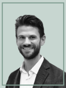 Professional headshot of a smiling man with dark hair and beard, wearing a blazer and white shirt against a pale green backdrop.