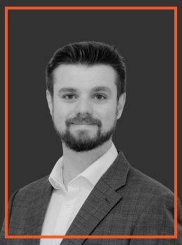 Professional headshot of a man with short dark hair and beard wearing a gray suit and white shirt against a dark background.
