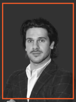 Head-and-shoulders portrait of a man with dark hair, wearing a checkered blazer and white shirt, looking at the camera.